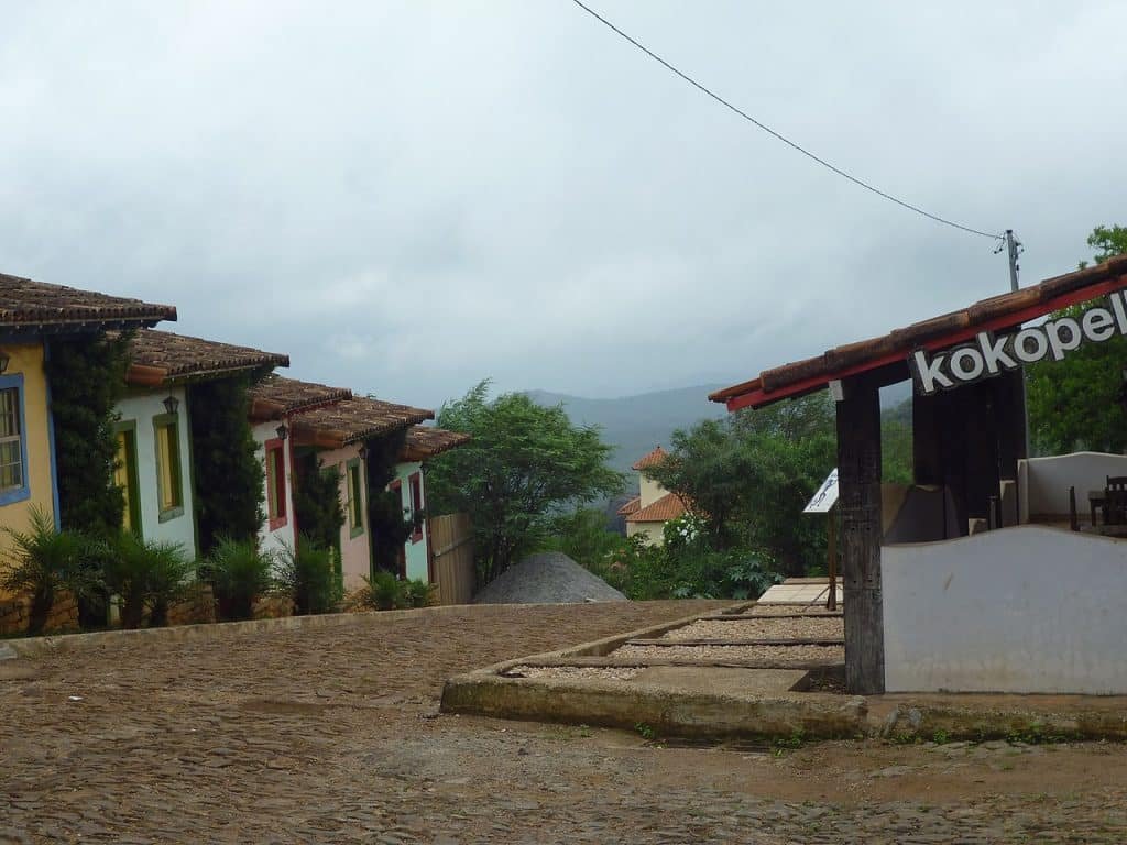 Rua de paralelepípedos ladeada por casas coloridas em um dia nublado. Uma placa de madeira diz "kokopelli". Árvores e colinas ao fundo.