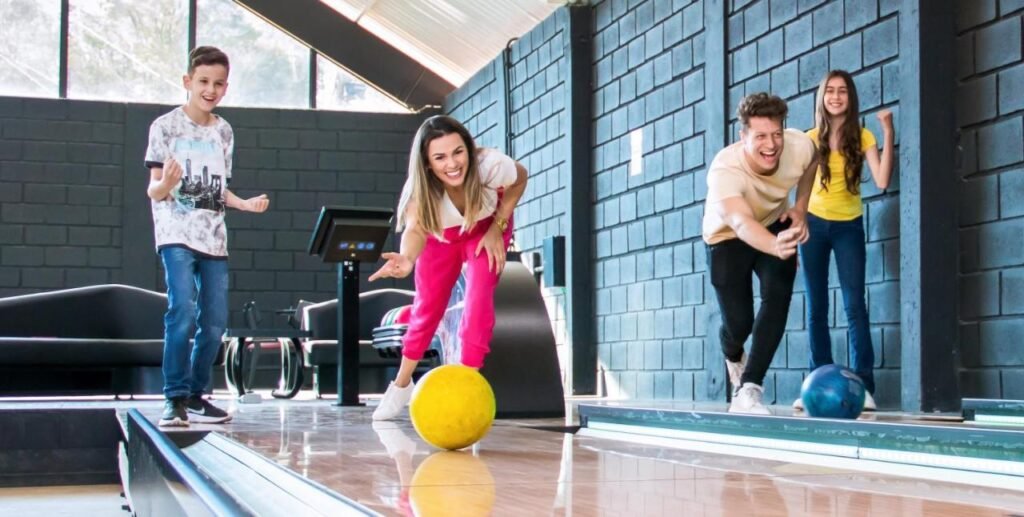 Dois adultos e duas crianças se divertem jogando boliche em uma pista coberta, sorrindo enquanto lançam as bolas nas pistas do Cassino All Inclusive Resort Poços de Caldas.