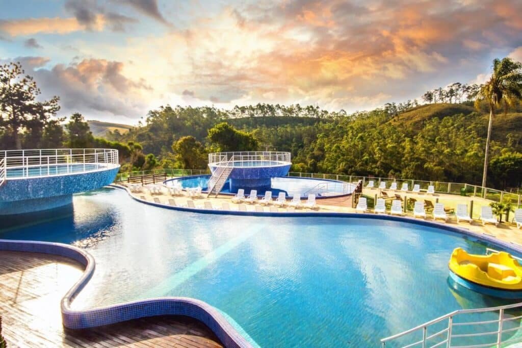 Uma grande piscina ao ar livre com uma plataforma circular, cercada por espreguiçadeiras e vegetação, sob um céu parcialmente nublado ao pôr do sol no Cassino All Inclusive Resort Poços de Caldas.