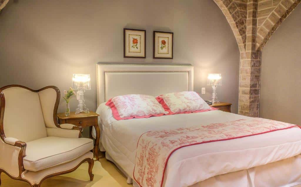 Cama de casal no Castello Benvenutti, uma das alternativas aos hotéis em Bento Gonçalves, com dois quadros sobre a cabeceira, mesinhas com abajur em cada lado da cama, uma poltrona e canteiros das paredes de tijolinhos