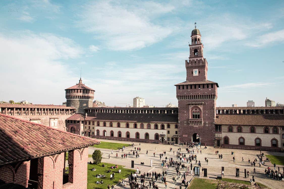 Lado de fora do Castello Sforzesco com uma torre central e um pátio, cercado por pessoas caminhando e se reunindo sob um céu azul claro.