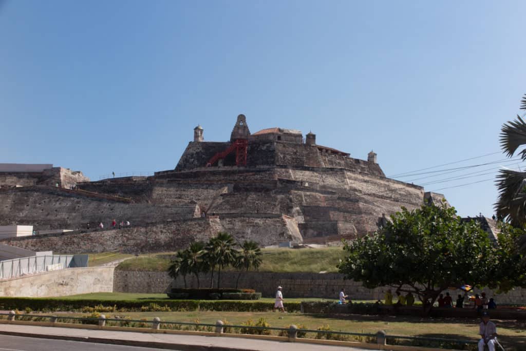 A imagem mostra uma fortaleza imponente feita de pedra, conhecida como Castillo San Felipe de Barajas, situada em um morro de Cartagena. A estrutura é composta por várias camadas de muralhas e torres defensivas, com uma rampa e escadas vermelhas visíveis, permitindo o acesso aos níveis superiores. Em primeiro plano, há uma área ajardinada com palmeiras e arbustos, onde algumas pessoas caminham ou descansam. O céu está claro e azul, indicando um dia ensolarado.