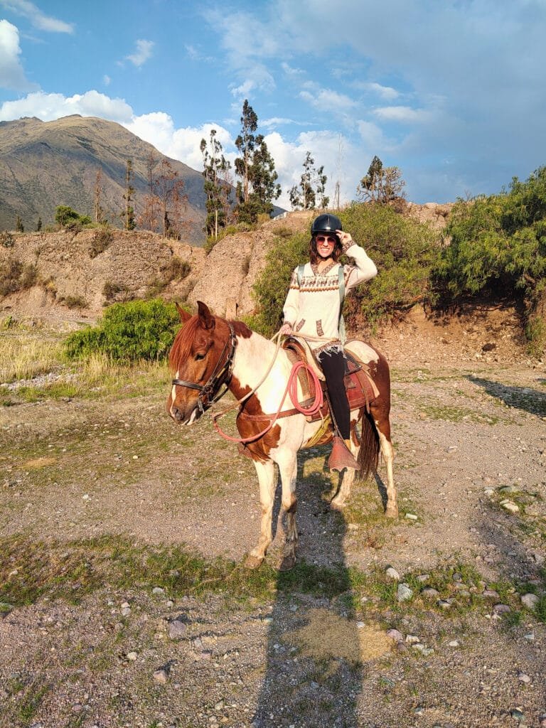 cavalo com pelagem marrom e branca ao centro da imagem, com várias montanhas ao fundo e um céu azul em contraste. Montada no cavalo, há uma mulher sorrindo, usando óculos escuros e capacete.