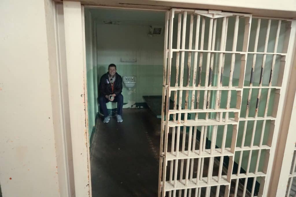 Homem sentado dentro de uma das celas da prisão de Alcatraz. O portão está aberto, a cama fica ao lado direito e uma pia no meio do ambiente, na parede do fundo.
