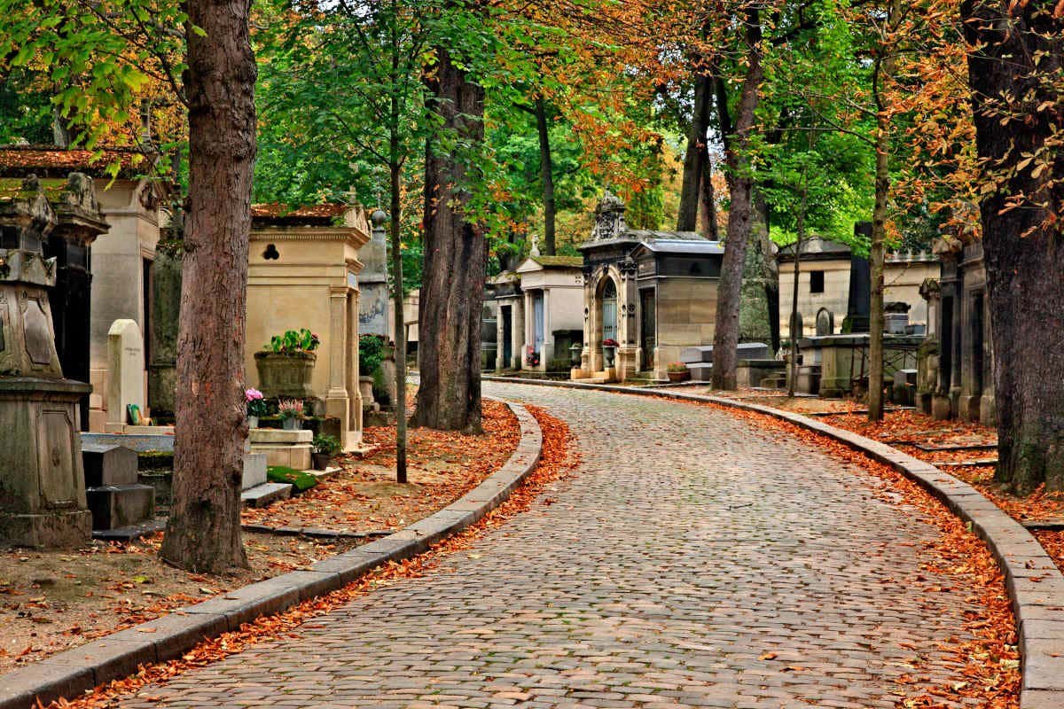 Um caminho de paralelepípedos curvo serpenteia por um cemitério com lápides ornamentadas e árvores de outono perdendo folhas coloridas.