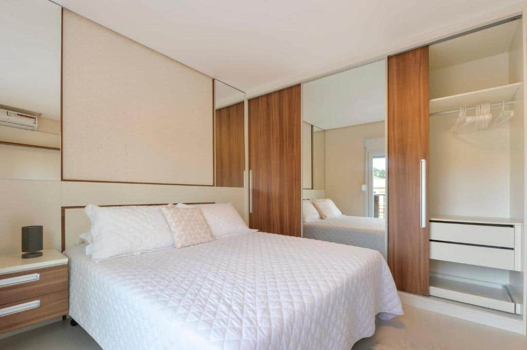 Quarto no Centro de Canela Apartamento Lindo. A cama box de casal está no meio de duas mesas de cabeceira com gaveta. Na direita da cama há um guarda-roupa bastante longo com espelho. Representa o post sobre airbnb em Canela.
