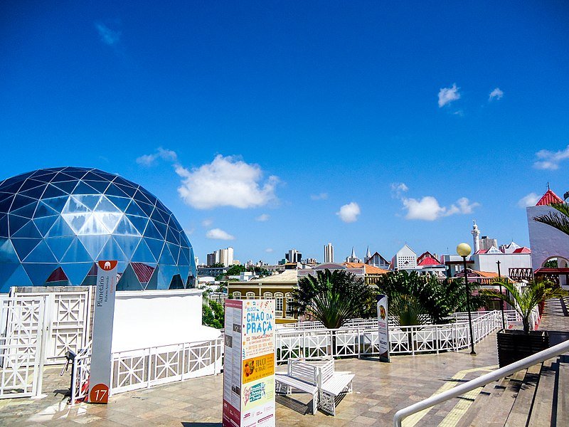 Foto do Centro Dragão do Mar de Arte e Cultura em Fortaleza. Está de dia e o céu possui poucas nuvens. Vemos um pátio com alguns cartazes informativos e plantas decorando. Na esquerda há um globo, que é um planetário do centro cultural. Bem no fundo da foto vemos alguns prédios e construções.