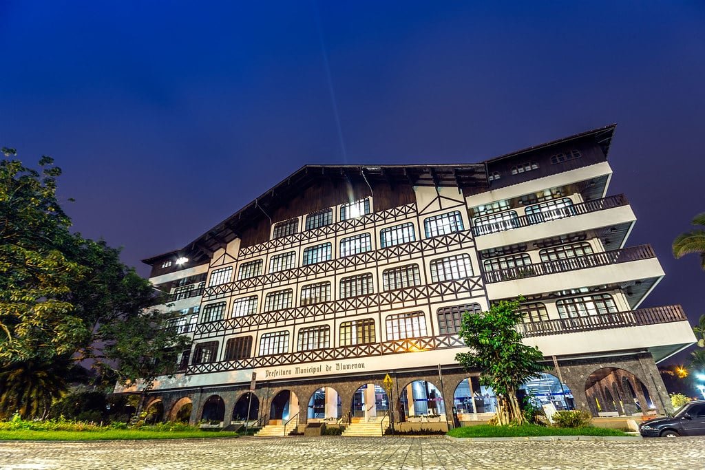 Prefeitura de Blumenau com várias sacadas, iluminado à noite. Árvores ladeiam a estrutura, e um caminho de paralelepípedos leva até ela. Imagem para ilustrar post sobre o que fazer em Blumenau.