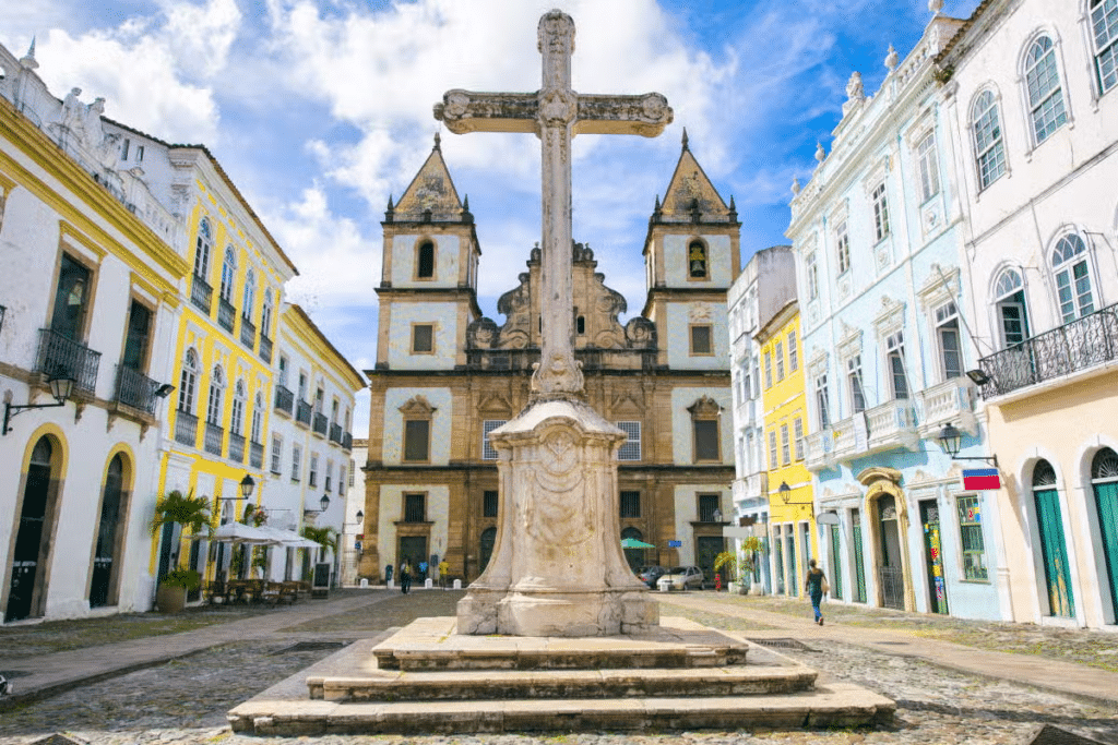 Uma cruz de pedra fica em uma praça do Centro Histórico de Salvador cercada por edifícios históricos coloridos e uma igreja com duas torres sineiras. Imagem para ilustrar post sobre o que fazer no feriado de Corpus Christi.