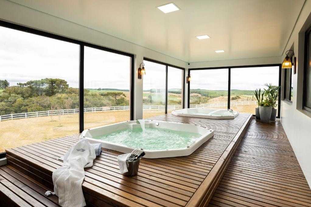 Uma sala de spa moderna com duas grandes banheiras de hidromassagem em plataformas de madeira, uma toalha e um balde de gelo. Grandes janelas oferecem vistas do campo.  Esse é o Cerro Azul Hotel Fazenda, um dos hotéis fazenda em Santa Catarina.
