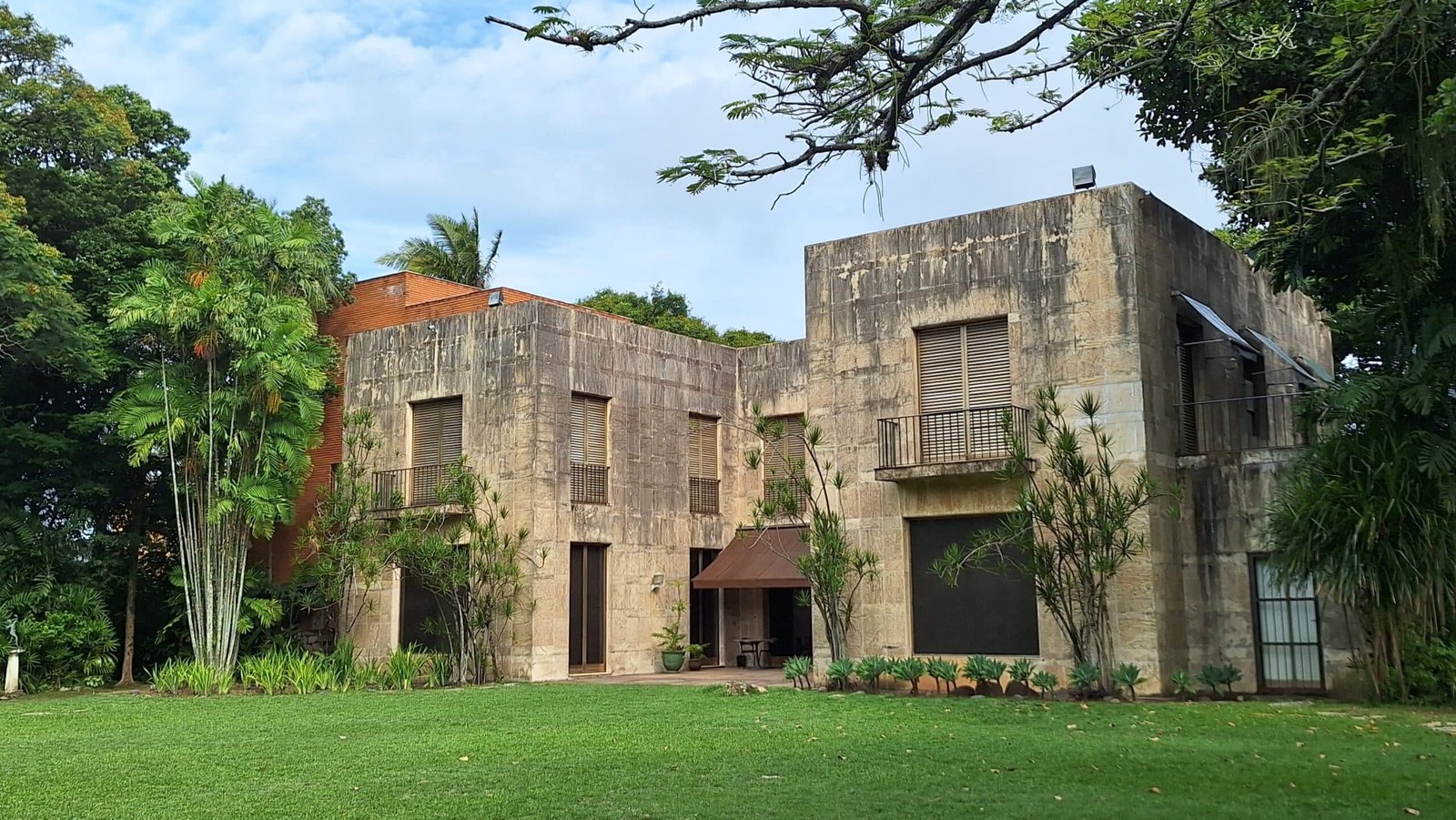 Casa de concreto com telhado plano, cercada por árvores e vegetação, com janelas retangulares e um pequeno pátio.