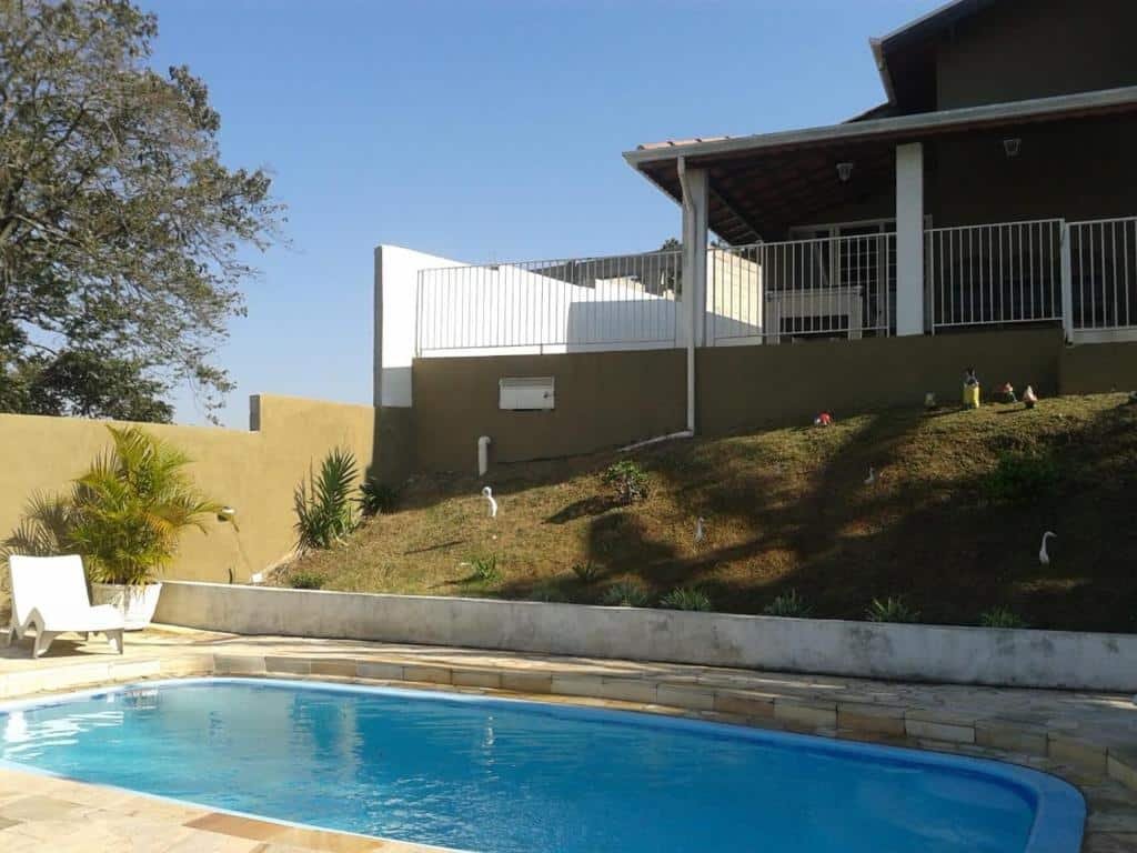 Uma piscina em um deck de pedra em Chácara Recanto Paraíso. Há uma espreguiçadeira no fundo à esquerda. Subindo um pequeno morro está a casa.