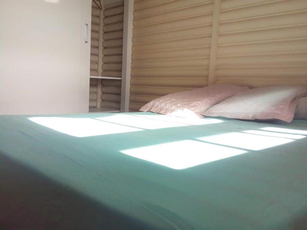 Quarto do Chalé Alpino mobiliado. A imagem mostra um quarto com uma cama coberta por um edredom verde. Há dois travesseiros cor-de-rosa sobre a cama. A luz do sol entra pelas persianas parcialmente fechadas da janela, criando padrões retangulares de luz na cama e nas paredes.