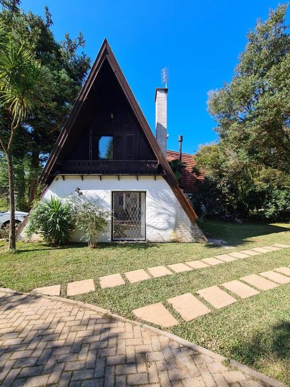 Foto do exterior do Chalé do Bosque. Sua estrutura é diferenciada, totalmente triangular. Há um gramado e um caminho de pedras na sua frente. O fundo possui algumas árvores e é possível ver outro chalé.
