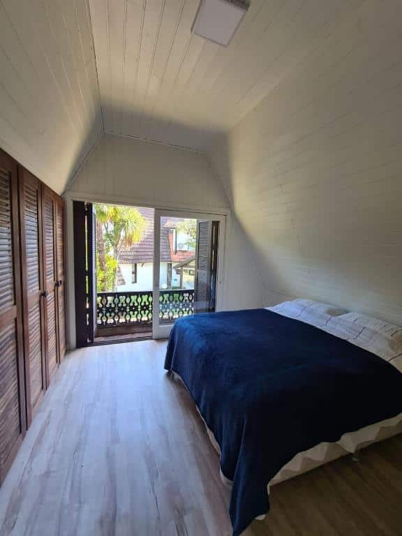 Foto do interior do Chalé do Bosque representando o post de chalés em Gramado. O quarto é novo e conta com uma cama de casal na direita, e na sua frente há uma parede com armários. O fundo da foto reserva uma varanda.