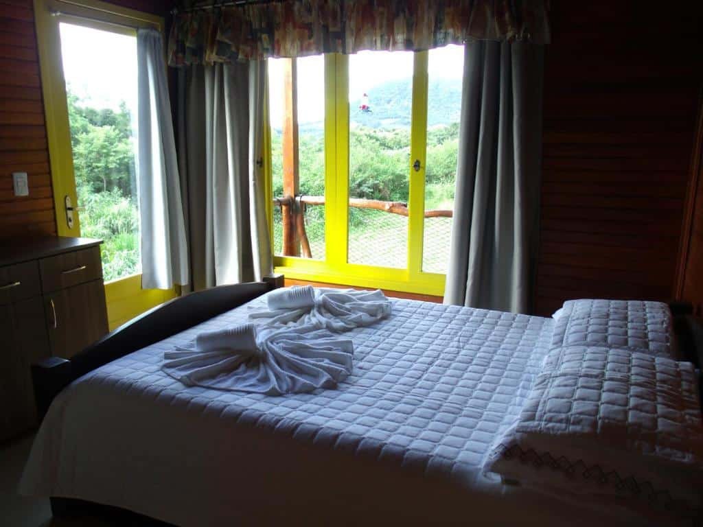 Foto do interior do Chalé dos Beija Flores representando o post de chalés em Gramado. Há uma cama box de casal na foto, e na sua frente há uma porta e janela com varanda e vista para a serra.