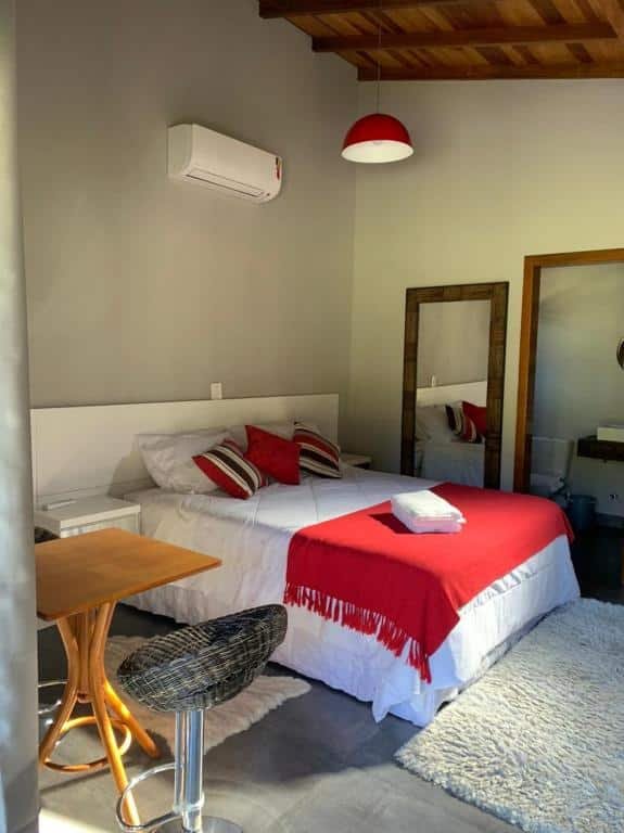 Foto do interior do Chalé dos Pássaros representando o post de chalés em Gramado. Há uma cama de casal no centro e um abajur no teto. Ao lado da cama de casal há duas escrivaninhas. No canto direito da foto se vê uma porta que leva ao banheiro.