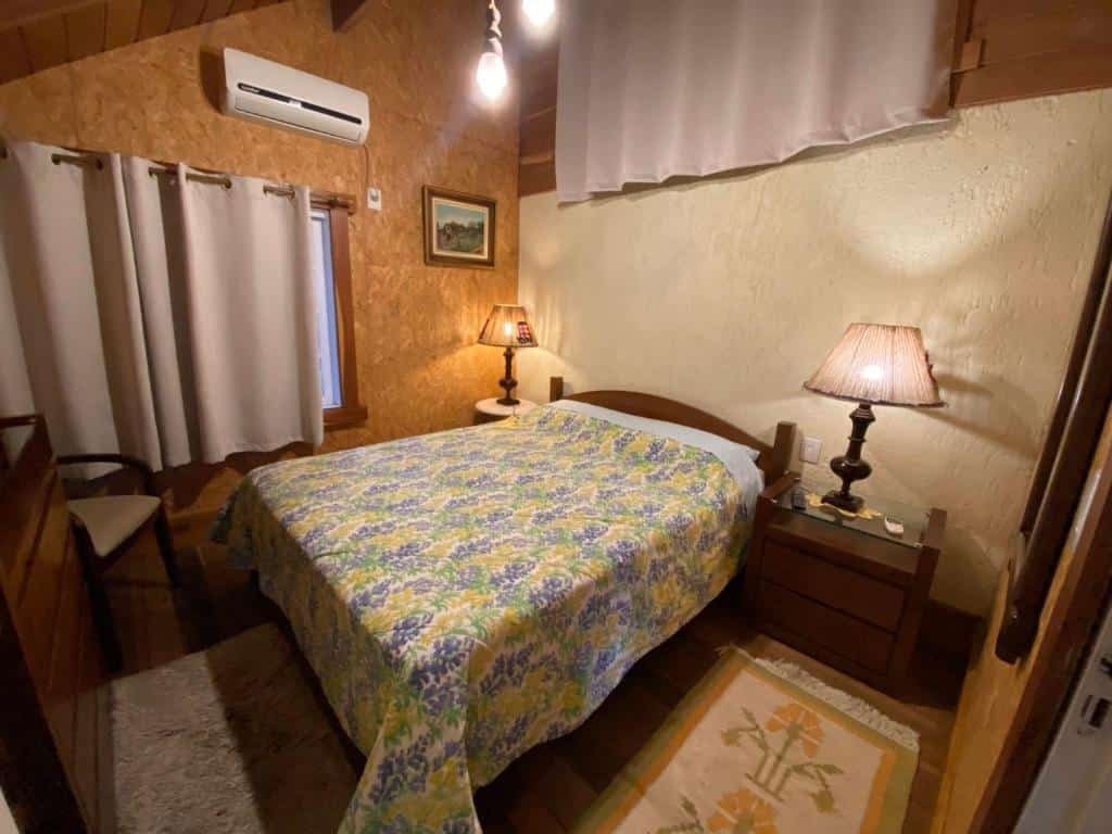 Quarto do Chalé Estilo Alemão. No centro, há uma cama de casal com uma colcha floral. Ao lado da cama, há mesas de cabeceira com abajures; um deles está aceso. Acima da cabeceira, há um quadro pendurado, e um ar-condicionado está instalado na parede alta. O quarto tem duas janelas com cortinas, uma delas parcialmente aberta, permitindo a entrada de luz natural. Um tapete felpudo está colocado aos pés da cama, adicionando um toque de conforto ao ambiente.