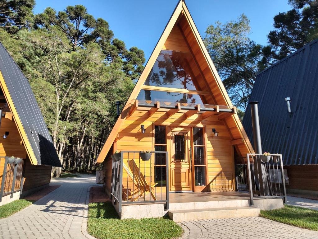 A imagem mostra uma cabana do Chalé Magia das Hortênsias moderna em estilo A-frame, com grandes janelas de vidro na fachada frontal, que permitem a entrada de muita luz natural. A cabana é construída com madeira de cor clara e tem um telhado inclinado acentuadamente, típico das estruturas em A-frame. A entrada possui uma pequena varanda com corrimão e degraus que levam a portas duplas. Ao redor da cabana, há altos pinheiros