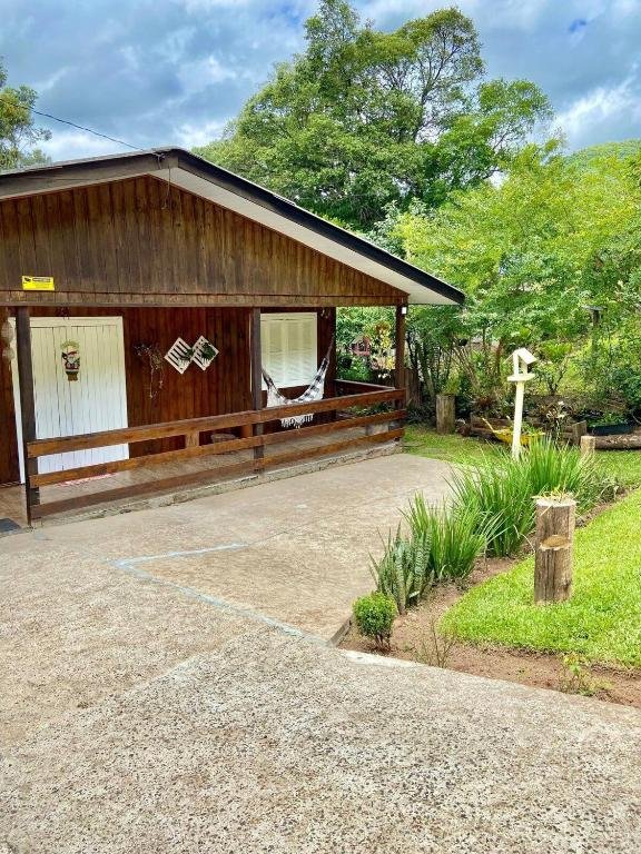 A foto é do Chalé Recanto dos Pássaros em Gramado e mostra um chalé de madeira com um telhado de duas águas e uma varanda coberta. Na frente do chalé, há duas janelas com cortinas brancas e uma porta branca visível no lado esquerdo. Em frente ao chalé, há um gramado bem cuidado com um caminho de concreto que leva à varanda. À direita do caminho, há uma pequena área de jardim com várias plantas e arbustos, além de um poste ou toco de madeira ornamental. Também há uma estrutura em forma de cruz no jardim à direita. O céu está parcialmente nublado, sugerindo que pode ser de manhã cedo ou fim de tarde