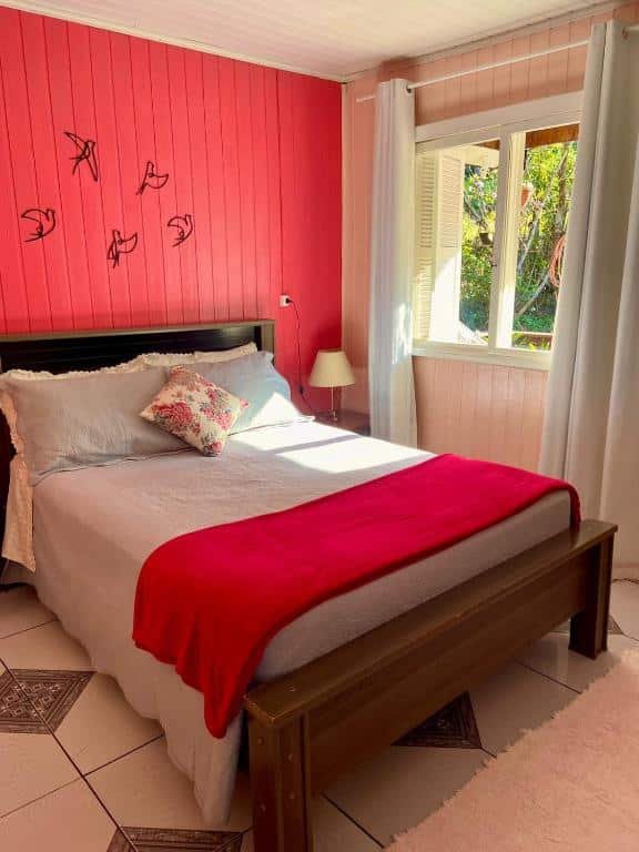 A foto é do Chalé Recanto dos Pássaros em Gramado e mostra um quarto acolhedor com uma parede de destaque vermelha vibrante, decorada com silhuetas de pássaros pretos. Há uma cama de casal com roupa de cama branca e uma manta vermelha aos pés. Um travesseiro com padrões florais está sobre a cama. À direita, há uma janela que permite a entrada de luz natural, e à esquerda, uma mesa de cabeceira de madeira com um abajur. O chão é de azulejo, e há um tapete rosa parcialmente sob a cama.