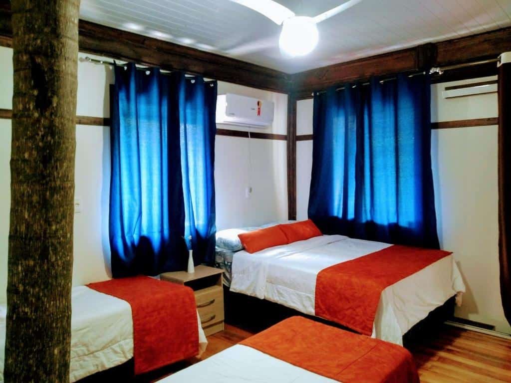 A foto mostra um quarto com três camas, cada uma coberta com colchas laranjas e um caminho de cama vermelho nos pés. Entre as camas, há um criado-mudo com uma pequena gaveta. O quarto tem paredes e piso de madeira. Um ventilador de teto branco está montado acima, e uma unidade de ar condicionado está instalada na parede à direita. Dois pilares de madeira natural também estão presentes no quarto, sugerindo um design rústico ou naturalista.