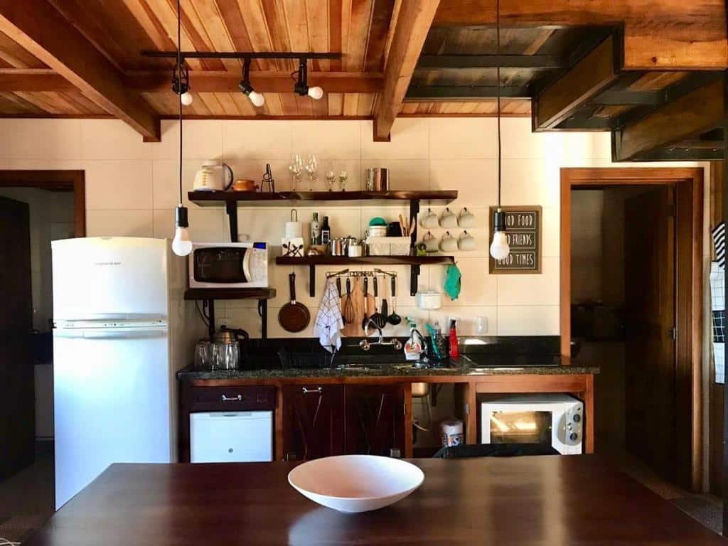 A foto mostra o interior de uma cozinha, provavelmente do Chalé Romântico em Gramado. A cozinha tem um estilo rústico, com vigas de madeira no teto e armários de madeira. Há um refrigerador, micro-ondas e um forno com fogão. Diversos utensílios de cozinha estão pendurados na parede, e há prateleiras acima do balcão exibindo copos e outros itens de cozinha. Uma grande tigela branca está colocada sobre o que parece ser uma mesa de jantar em primeiro plano. O ambiente é aconchegante e convidativo, ideal para quem busca uma estadia charmosa em Gramado.