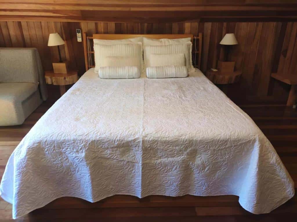 A foto mostra um quarto aconchegante com paredes e piso de madeira, típico de um chalé romântico em Gramado. A cama está bem arrumada com uma colcha branca texturizada e dois travesseiros. Há dois abajures, um de cada lado da cama, criando uma iluminação suave e acolhedora. À esquerda da cama, há uma poltrona com uma almofada clara, adicionando um toque extra de conforto ao ambiente. A atmosfera é intimista e convidativa, perfeita para uma estadia romântica. 