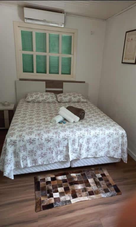 O quarto do Chalé São Rafael parece ser bastante acolhedor e charmoso. Ele possui uma cama grande com uma colcha de estampa floral, e um cabeceira de madeira. Acima da cama, há um quadro decorativo na parede. A janela, localizada acima da cama, tem persianas verdes, que adicionam um toque de cor ao ambiente. No chão de madeira, aos pés da cama, há um tapete xadrez em tons de marrom e bege, que complementa a decoração rústica do quarto.