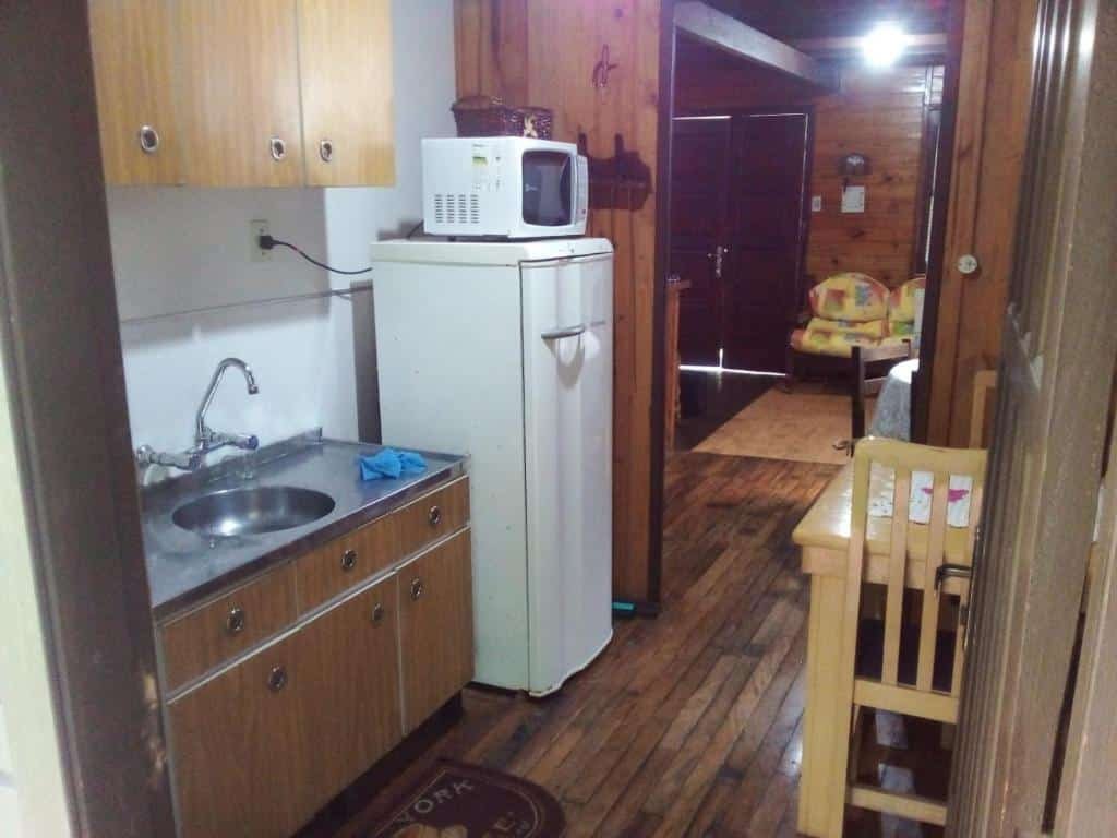 A imagem mostra uma vista interior de uma cozinha do chalé três quartos em Gramado que leva a outro cômodo. À esquerda, há um balcão de cozinha com uma pia e armários acima e abaixo. Um micro-ondas está sobre o balcão, e uma geladeira está ao lado. No primeiro plano à direita, há uma cadeira de madeira, e além dela, uma porta aberta leva a outro cômodo onde é visível um assento de balanço infantil. O piso parece ser de madeira em toda a extensão, e há um tapete com texto em frente à pia.