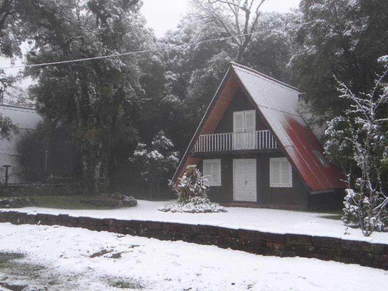 A foto mostra uma chalé de três quatros em Gramado com dois andares com um telhado vermelho inclinado, paredes brancas e uma varanda no andar superior. O chão está coberto de neve, e há árvores parcialmente visíveis através da névoa ao fundo. A cena captura um ambiente sereno de inverno.