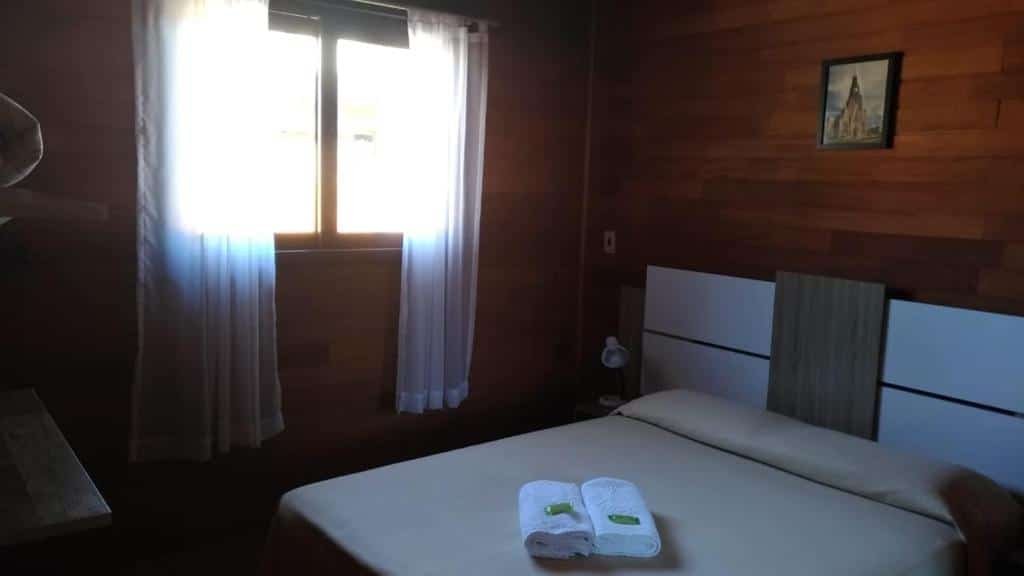 Quarto no Chalés Araucárias da Serra. A cama de casal está na direita, e atrás dela há uma cabeceira com abajur. Do seu lado esquerdo há uma janela. As paredes são de madeira. Representa o post sobre airbnb em Canela.