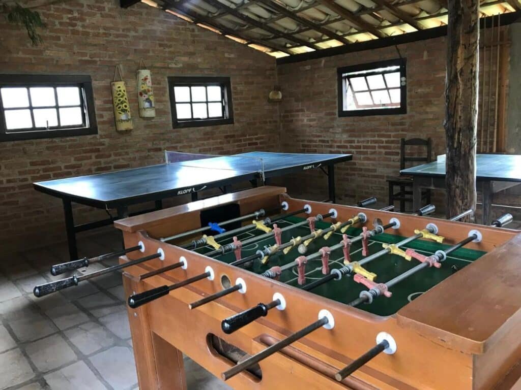A imagem mostra uma sala de jogos com paredes de tijolos aparentes e telhado de telhas de barro. No centro, há uma mesa de pebolim em madeira com bonecos coloridos prontos para o jogo. Ao fundo, há duas mesas de pingue-pongue lado a lado. O ambiente é iluminado por pequenas janelas de madeira que deixam a luz natural entrar. A decoração é simples, com alguns itens pendurados nas paredes. O espaço parece ser rústico e descontraído, ideal para momentos de lazer. Esse é o Chalés de Minas Hotel Fazenda, um dos hotéis-fazenda em Minas Gerais.