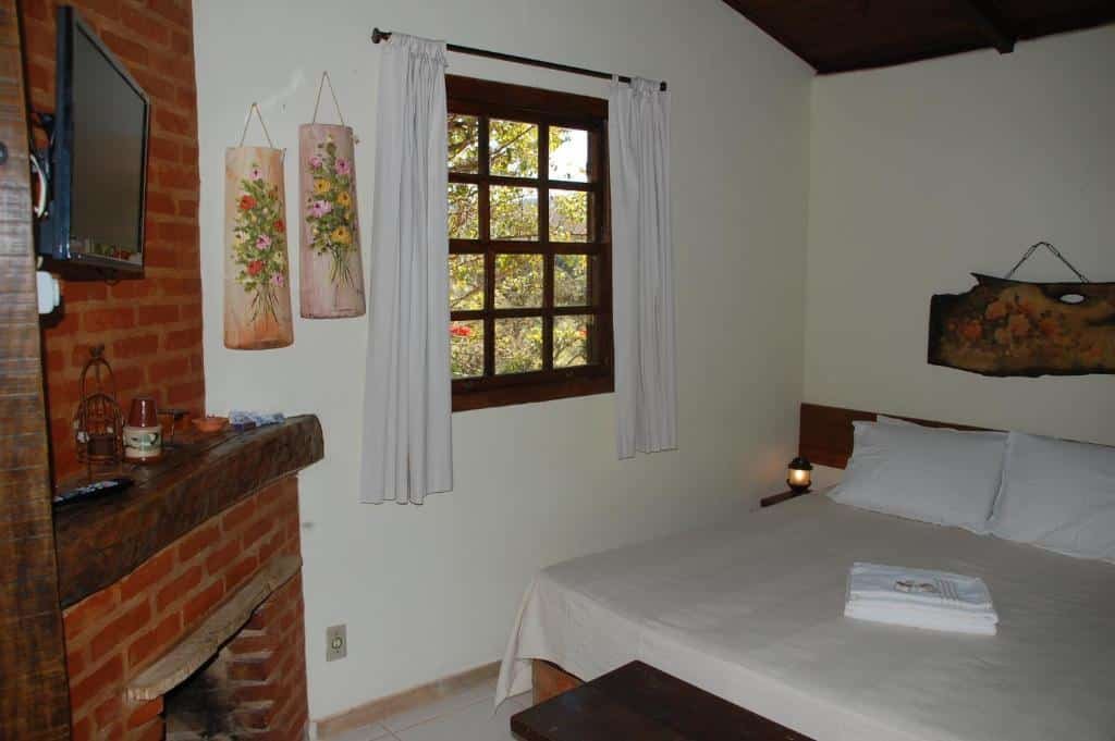 A imagem mostra um quarto aconchegante com decoração rústica no Chalés de Minas Hotel Fazenda. À esquerda, há uma lareira de tijolos com uma prateleira de madeira acima, decorada com pequenos objetos e um controle remoto. Ao lado, uma janela de madeira com cortinas brancas deixa entrar luz natural. A cama de casal está coberta com roupa de cama branca, e sobre ela há toalhas dobradas. Na parede acima da cama, há um enfeite de madeira com uma pintura. O ambiente parece acolhedor e tranquilo, ideal para descansar em um lugar campestre.