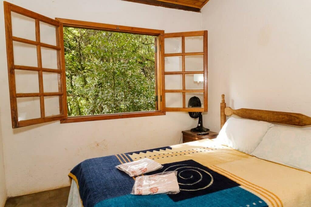 Quarto da Chalés do Beto, com uma cama de casal e duas toalhas dobradas dentro de um plástico em cima. Ao lado, há uma escrivaninha com ventilador em cima e uma janela mostrando folhas verdes da natureza