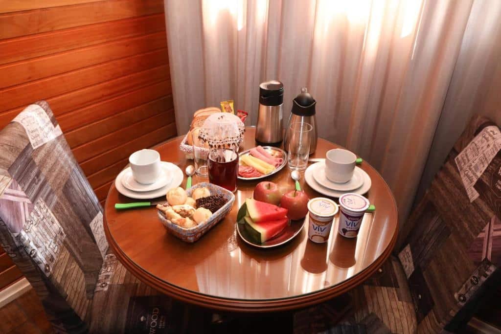 Uma mesa de café da manhã dentro do quarto dos chalés Poncho Serrano. Está com várias frutas, iogurtes, pães, frios, café e xícaras e talheres. Há dois lugares na mesa.