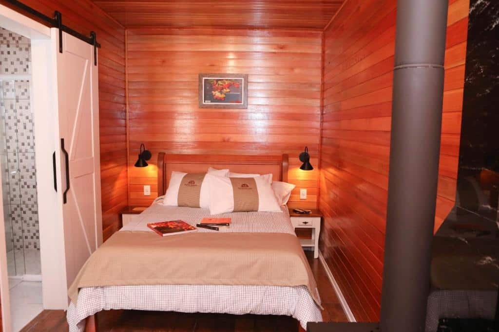 Foto do interior dos Chalés Poncho Serrano representando o post de chalés em Gramado. Há uma cama de casal no centro. Há dois criados-mudos com abajur que a ladeiam e uma porta está aberta à sua esquerda.