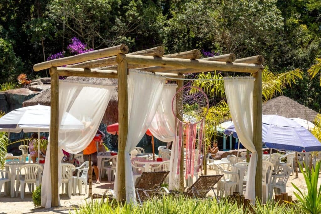 A imagem mostra uma área ao ar livre com uma estrutura de madeira tipo pergolado, coberta por cortinas brancas esvoaçantes, criando uma atmosfera relaxante nos Chalés Praia de Minas. Há cadeiras de praia de madeira embaixo do pergolado, e um filtro dos sonhos colorido pendurado em um dos pilares, adicionando um toque decorativo. Ao fundo, mesas e cadeiras brancas de plástico estão dispostas sob grandes guarda-sóis coloridos, oferecendo sombra para as pessoas que relaxam no local. A vegetação ao redor é densa, com árvores e plantas, completando o ambiente natural.