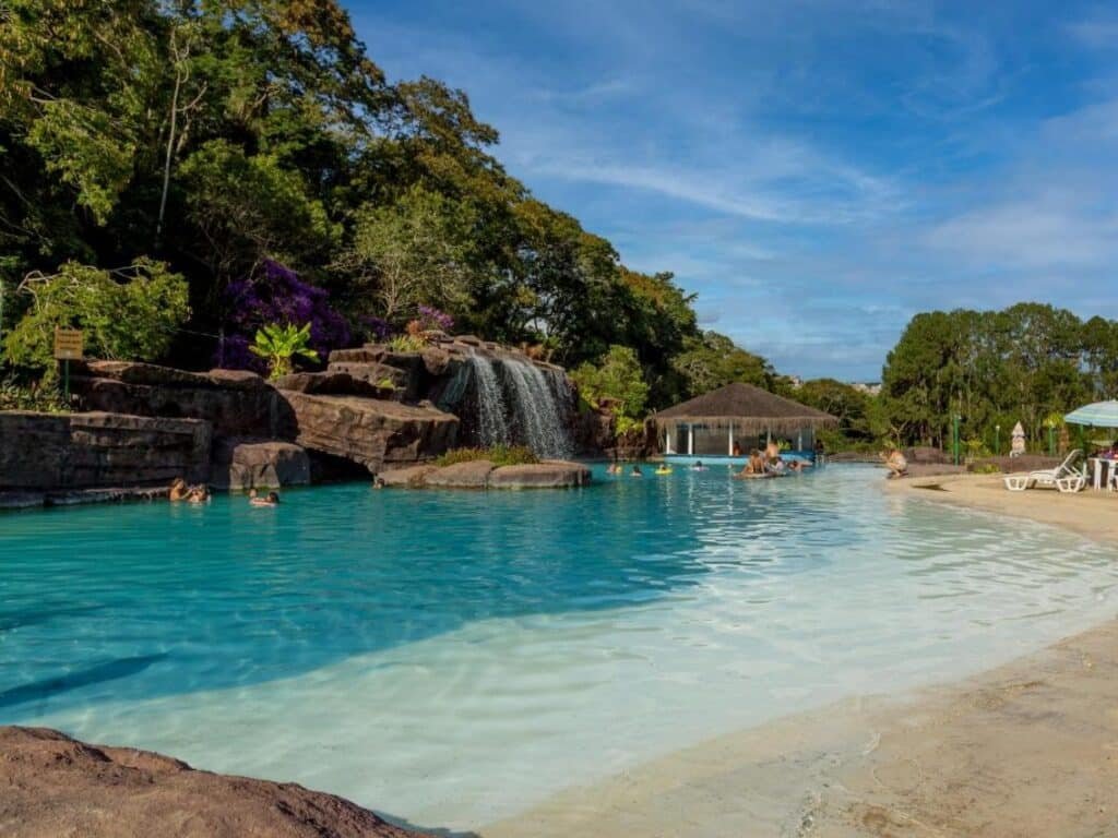 A imagem mostra uma piscina ampla nos Chalés Praia de Minas cercada por vegetação. No centro da piscina, há uma pequena cachoeira artificial caindo de uma formação rochosa, criando um efeito relaxante. Algumas pessoas estão nadando na água clara e azul, enquanto outras estão sentadas nas espreguiçadeiras à beira da piscina. Ao fundo, uma estrutura coberta de palha serve de bar molhado, onde mais pessoas se reúnem. O céu está claro, com poucas nuvens, e a paisagem é tranquila, ideal para relaxamento. Esse local é um dos hotéis-fazenda em Minas Gerais.