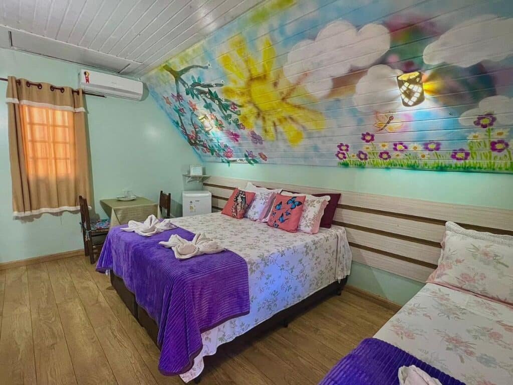 A imagem mostra um quarto de acolhedor nos Chalés Praia de Minas, com uma cama de casal e uma cama de solteiro, ambas cobertas por colchas florais e mantas roxas. Acima das camas, o teto inclinado é decorado com uma pintura vibrante de céu azul, nuvens, sol brilhante e flores coloridas. Há um pequeno frigobar ao lado da cama, uma mesa com cadeiras próximas à janela, que tem cortinas marrons, e um ar-condicionado fixado na parede. O ambiente é alegre e decorado com um toque artístico e confortável.