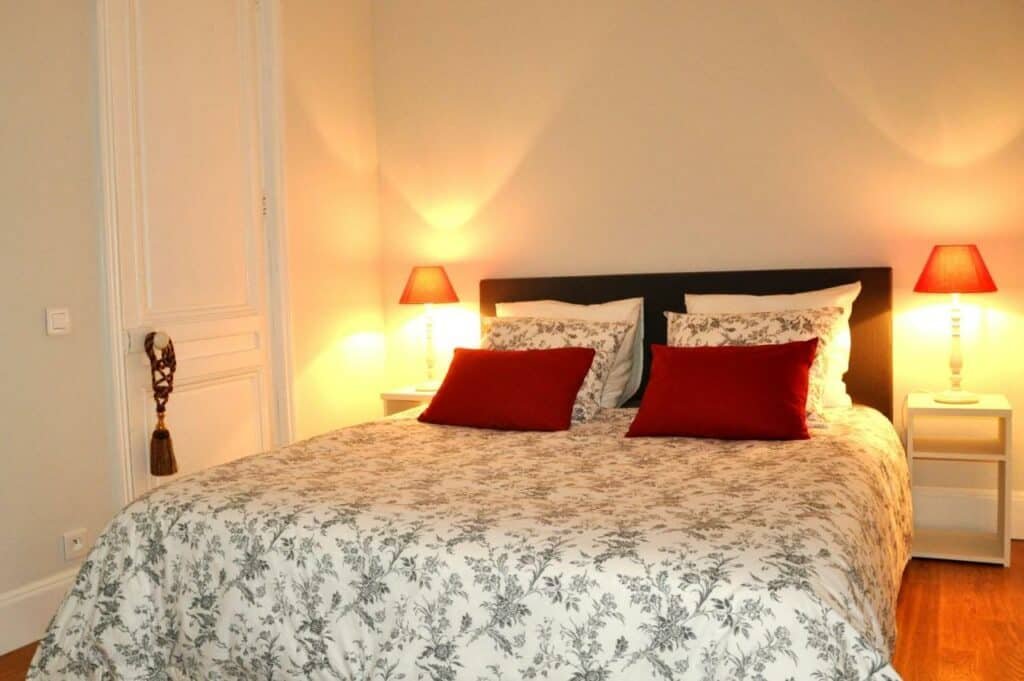 Quarto do Chambre De Flore. Uma cama de casal com almofadas tem mesinhas de cabeceira e abajures dos dois lados, e há uma porta na lateral esquerda.