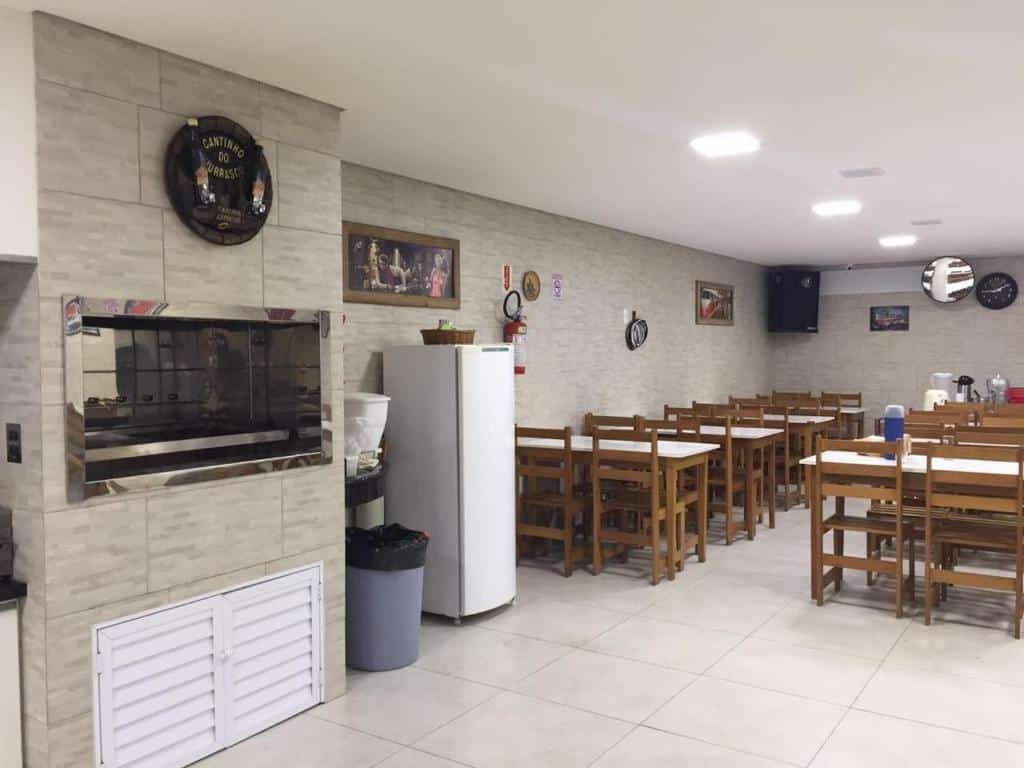 Cozinha do Chemin Suites com várias mesas com cadeiras, uma geladeira branca e um forno industrial no lado direito para ilustrar as pousadas em Balneário Camboriú