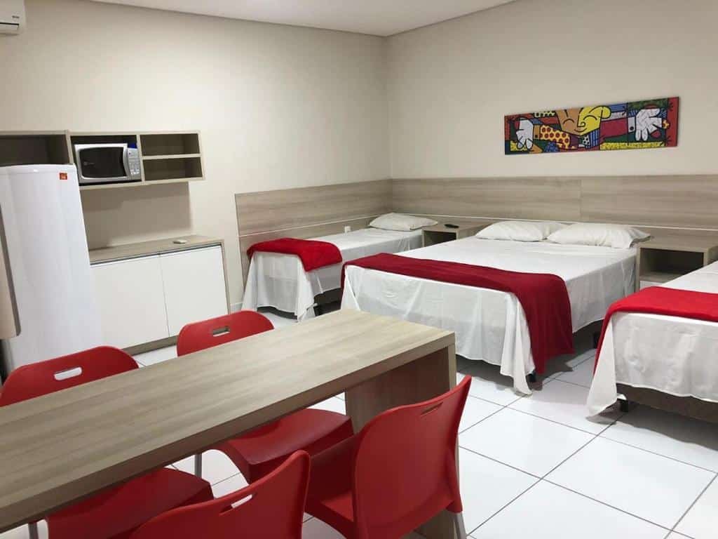 Quarto Queen Deluxe da Chemin Suites, com uma cama de casal e duas de solteiro, uma mesa de madeira com cadeiras vermelhas em volta e uma cozinha compacta com micro-ondas e geladeira ao lado