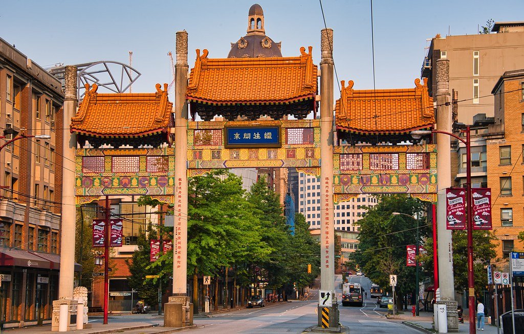Portão ornamentado com estilo arquitetônico chinês em um ambiente urbano, cercado por edifícios e árvores, sob um céu claro.