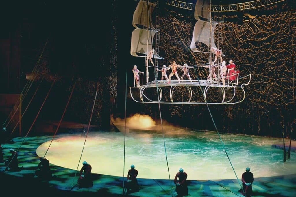 A imagem mostra uma das cenas do espetáculo "O", do Cirque du Soleil. Vários acrobatas estão suspensos em uma estrutura que imita um navio. Abaixo deles há uma piscina com fumaça e outros acrobatas ao redor. A foto está na lista de o que fazer em Las Vegas.