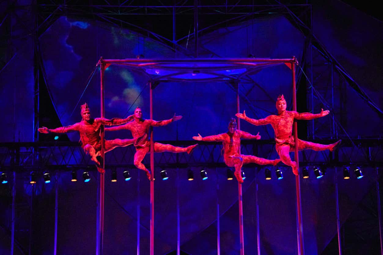 Quatro trapezistas do espetáculo Mystère, no Cirque du Soleil em Las Vegas. Eles estão em trajes vermelhos e realizam movimentos sincronizados em postes verticais sob iluminação colorida do palco.