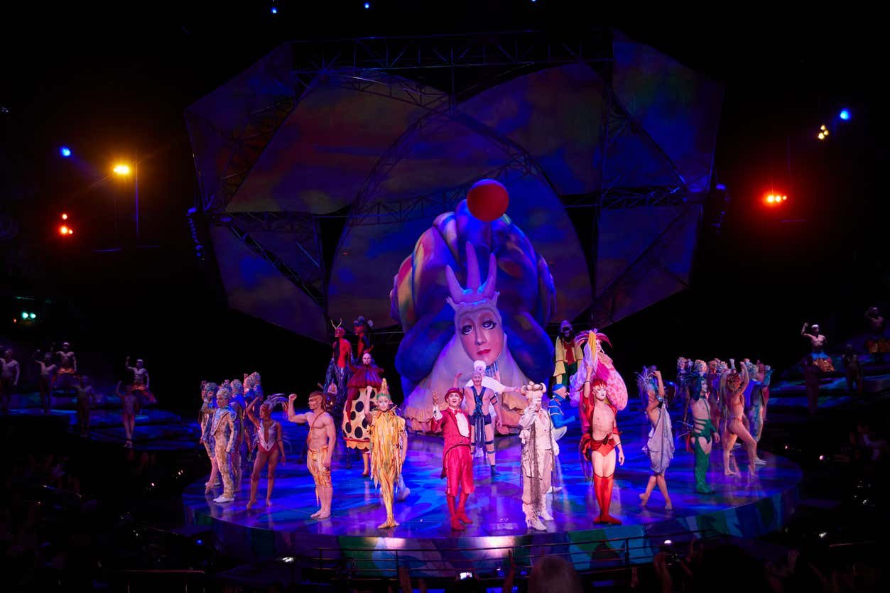 Artistas em trajes coloridos do espetáculo Mystère, no Cirque du Soleil em Las Vegas, em um palco bem iluminado com um grande cenário abstrato com um rosto e uma esfera vermelha.