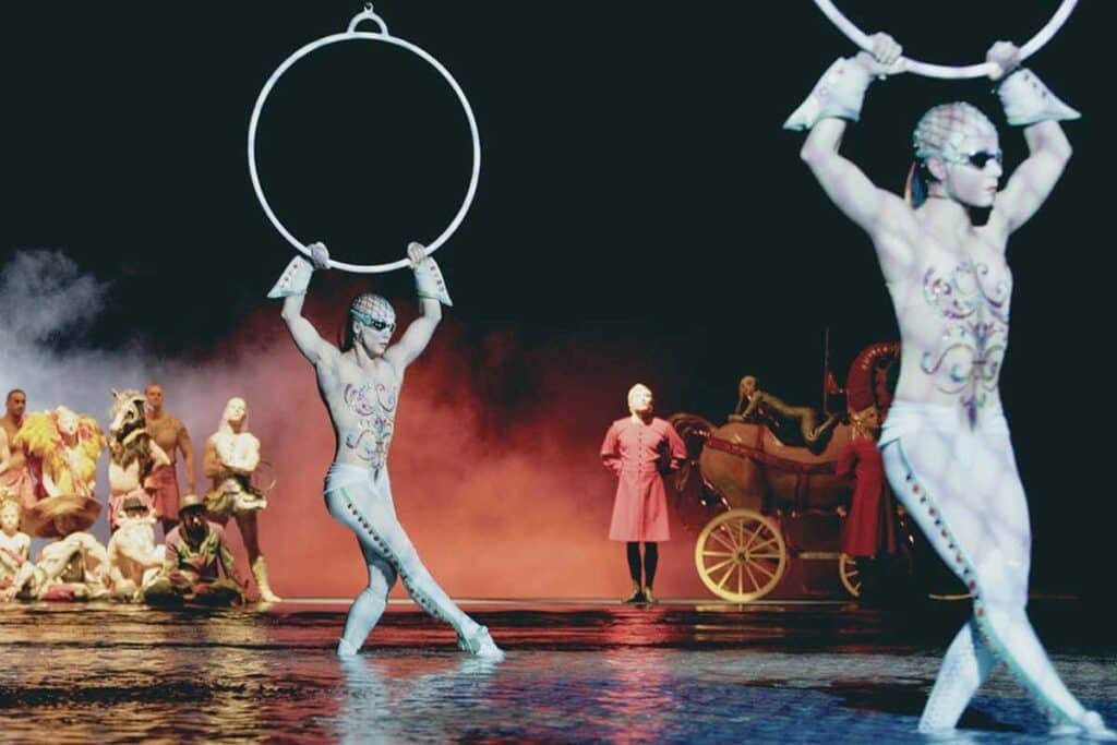 Artistas do espetáculo "O", do Cirque du Soleil em Las Vegas, em trajes estampados com anéis no palco, outros no fundo em trajes coloridos e uma carruagem vermelha.