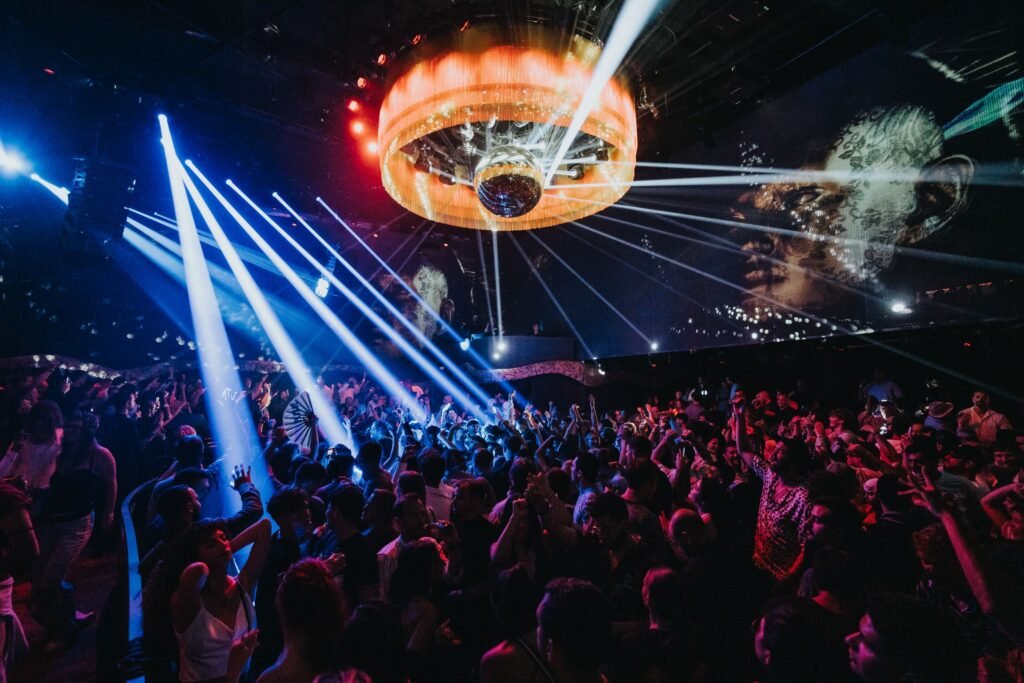 Uma multidão animada dança sob luzes coloridas e uma bola de discoteca em um ambiente escuro de boate, com imagens projetadas nas paredes. Esse é o Club Chinois, uma das festas em Ibiza.