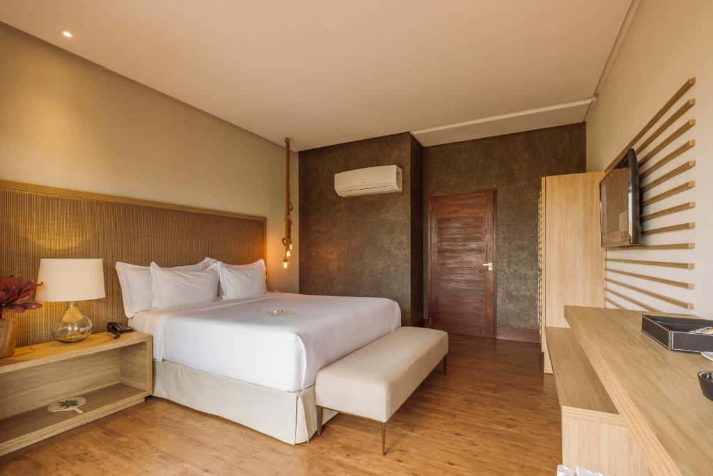 Quarto de casal na Colina Pousada SPA, com ar-condicionado, mesa de cabeceira com flores e abajur, cama ampla, recamier aos pés da cama, e TV na parede